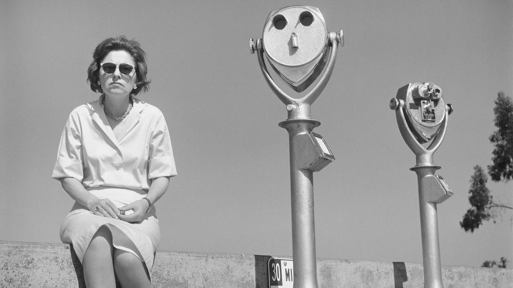 black and white photo of a woman sitting next to two coin-operated viewers; the viewer next to her echoes her expression