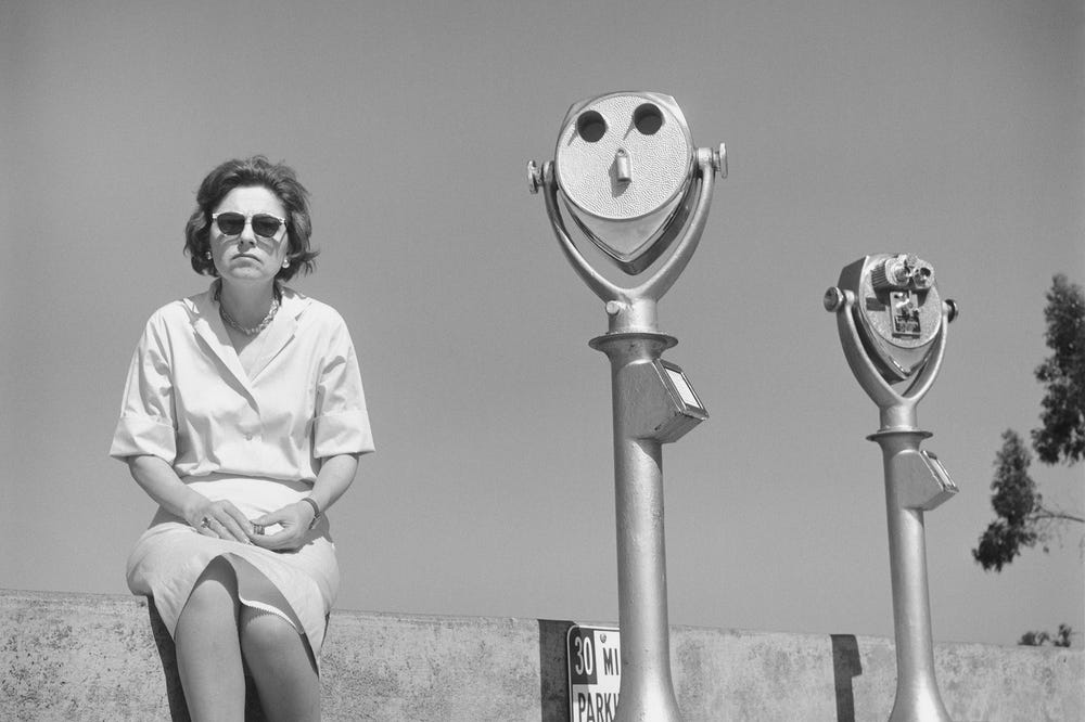 black and white photo of a woman sitting next to two coin-operated viewers; the viewer next to her echoes her expression