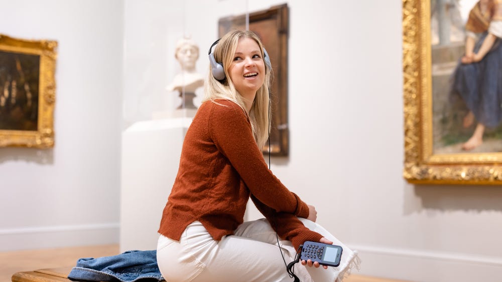 a young woman with an audio tour and headphones sits on a bench, looking back over her shoulder
