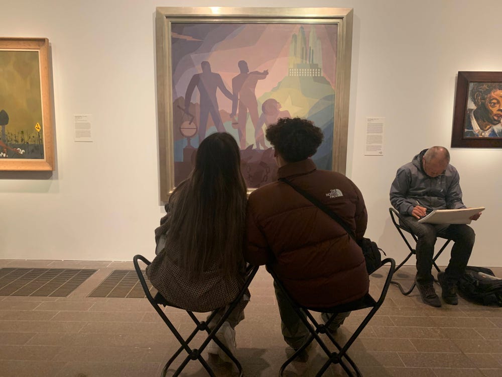 A seated couple, viewed from the back, looking at a painting of two large purple figures pointing toward a factory on a hill. Another man sketches next to the painting.