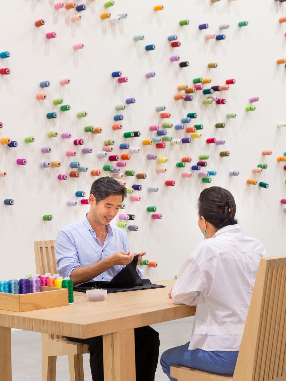 People mending cloth at a table in front of a wall covered in spools of thread