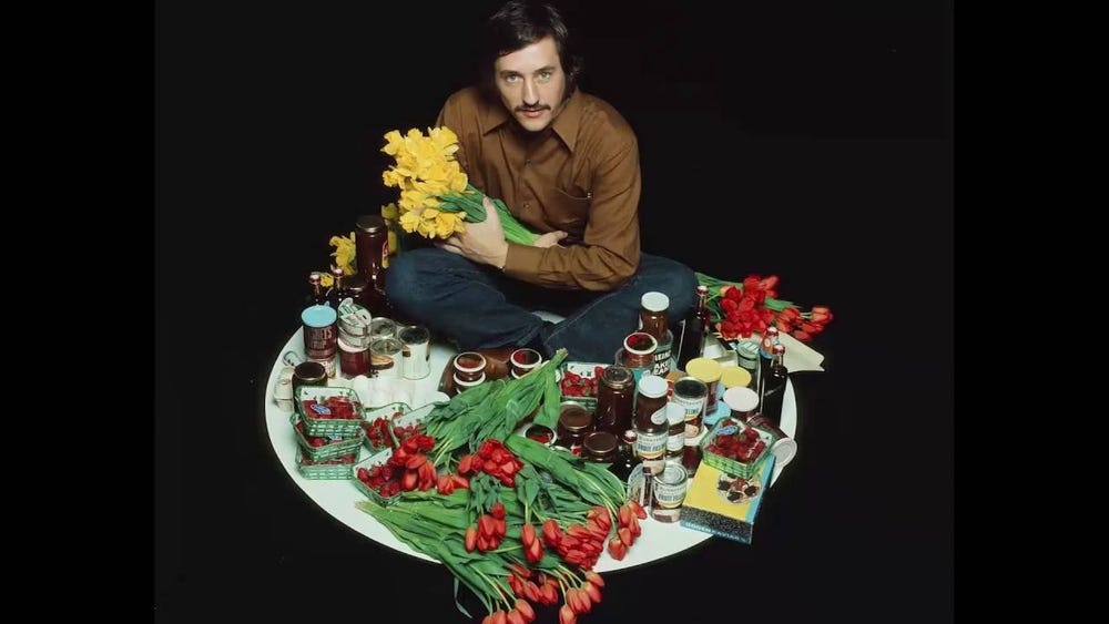 Ed Ruscha sitting on the ground with various foods and flowers