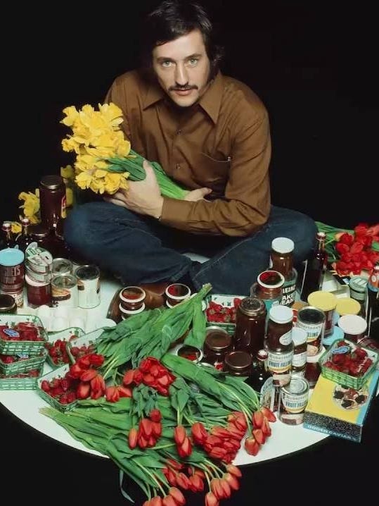 Ed Ruscha sitting on the ground with various foods and flowers
