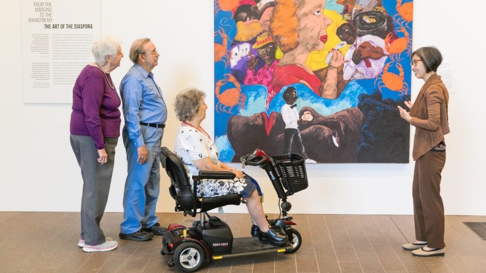 woman in a motorized scooter and two other patrons and a docent front of a painting