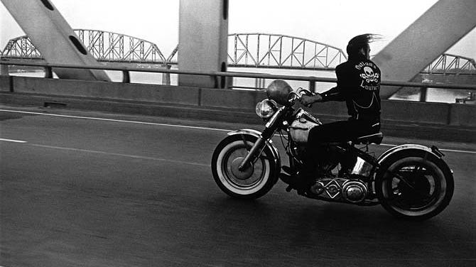 Black and white photo of man on motorcycle crossing bridge.