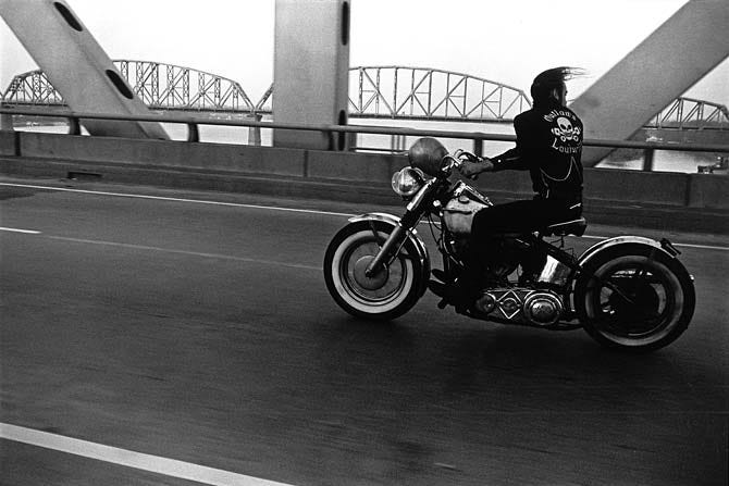 Black and white photo of man on motorcycle crossing bridge.