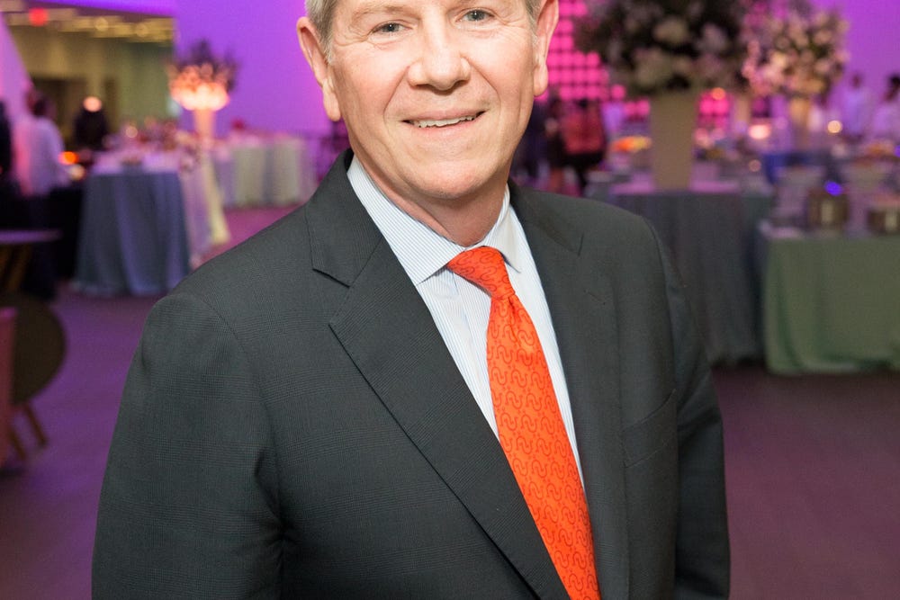 Photograph portrait of man wearing black suit and orange tie in front of purple background.