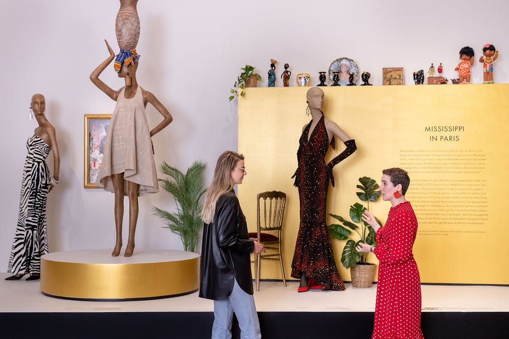 two women speaking to one another in front of an exhibition installation of Patrick Kelly