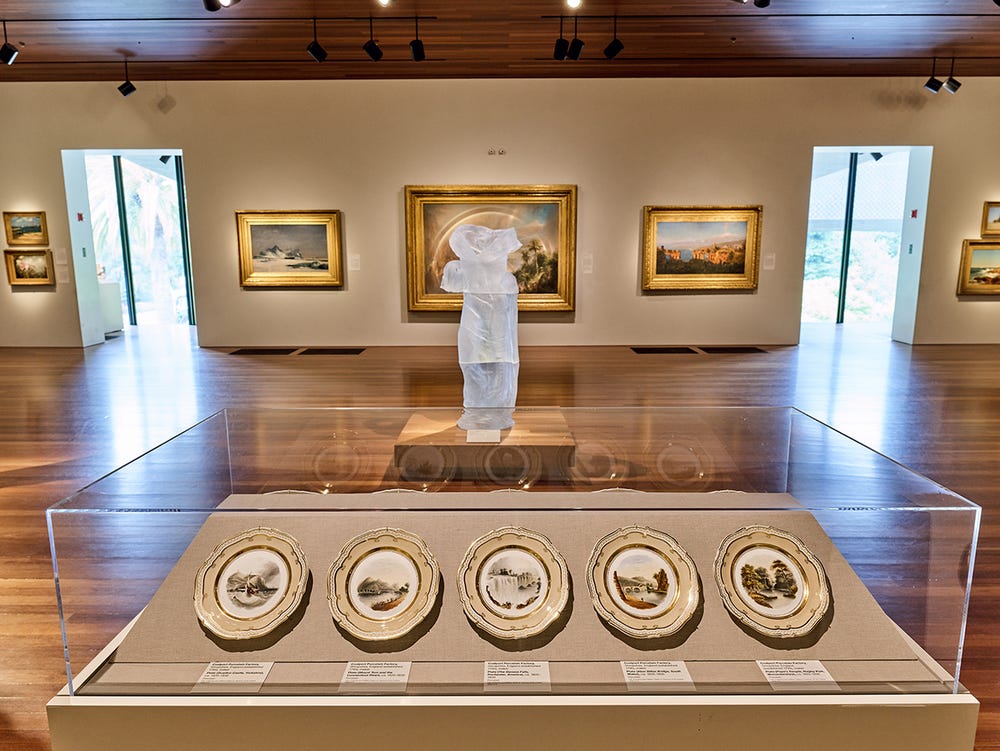 a gallery displaying a case with plates in front of a translucent sculpture and paintings