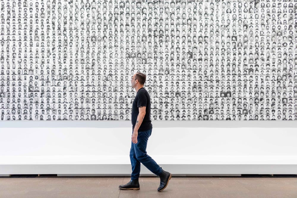 a man walking in front of an artwork featuring black and white headshots with the eyes blocked