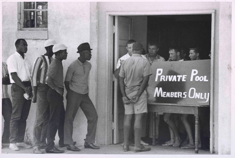 Black men line up at an entrance to a swimming pool, with white men behind a sign reading “Private pool, members only”