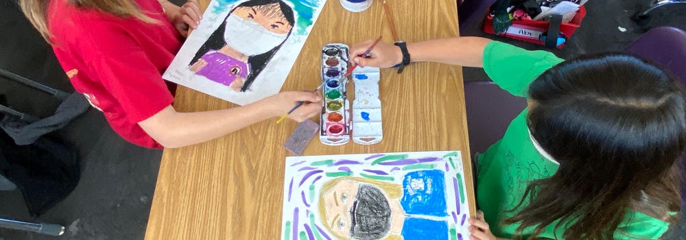 Overhead shot of two kids dipping paintbrushes into watercolor as they make portraits