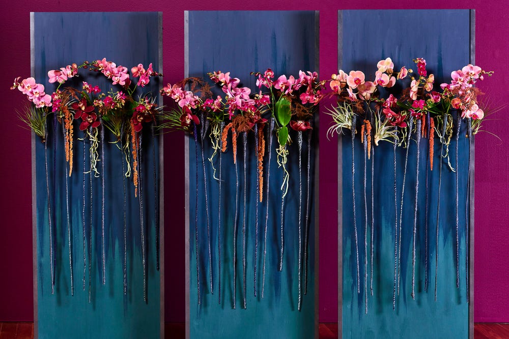 pink and red flowers with long stems hanging against three blue panels in front of a pink wall
