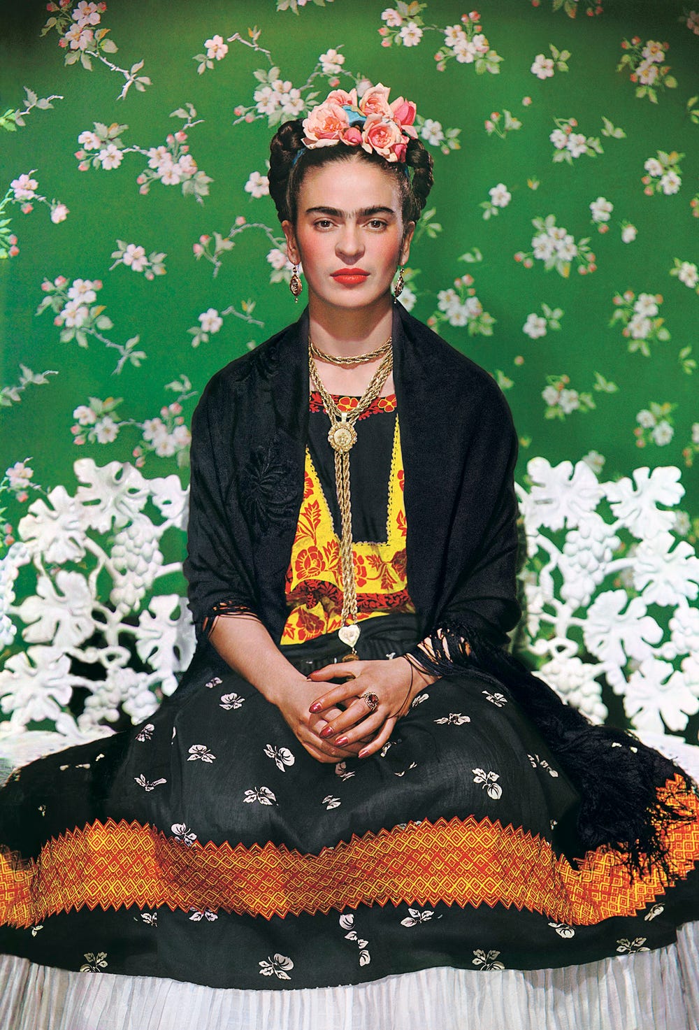 photo of a woman with dark hair and full eyebrows sitting on a white bench in front of a green floral background