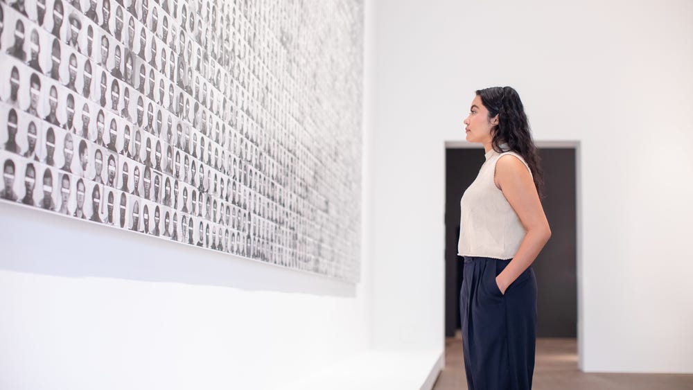 a woman looking at an artwork consisting of black and white headshots with eyes blocked out