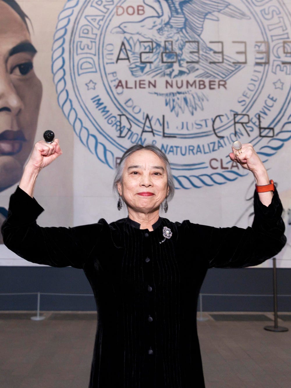 Hung Liu standing in front of her large-scale artwork of an Identification document with her biceps flexed.