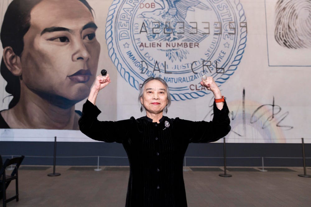 Hung Liu standing in front of her large-scale artwork of an Identification document with her biceps flexed.