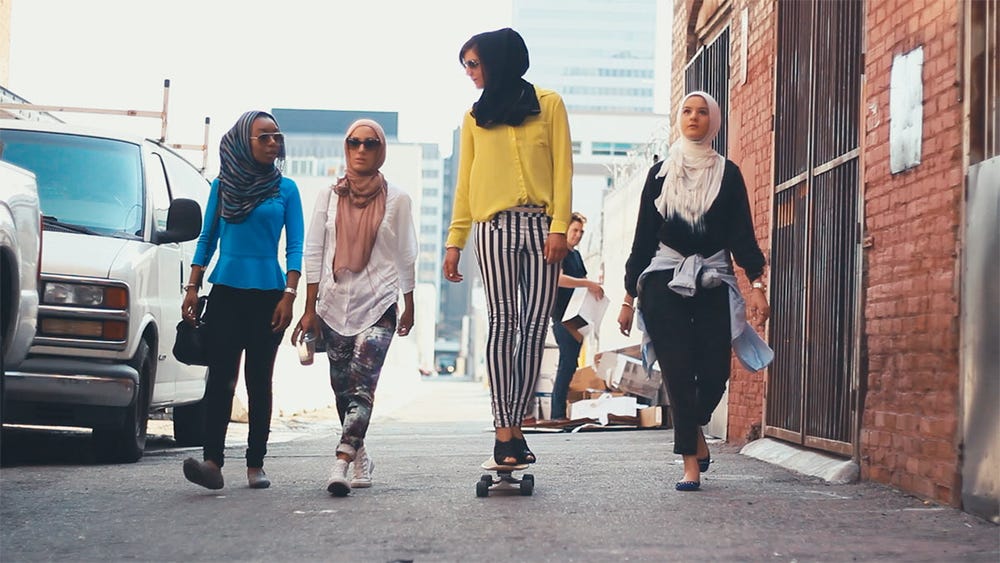 video still with four women with head coverings walking down the street, one on a skateboard