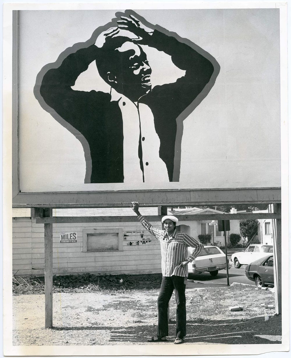 Cleveland Bellow photographed standing under his billboard, showing a Black boy with his hands raised above his head
