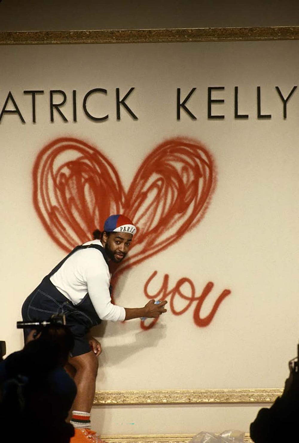 Patrick Kelly posing with spray paint in front of a sign that reads "Patrick Kelly [heart] you"