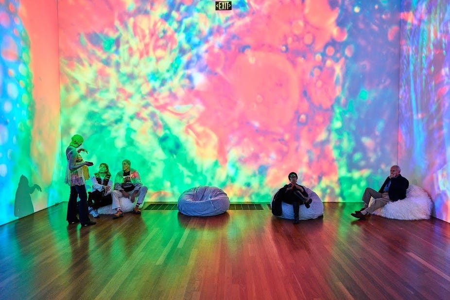 Photo of patrons sitting on floor of art gallery surrounded by colorful walls.