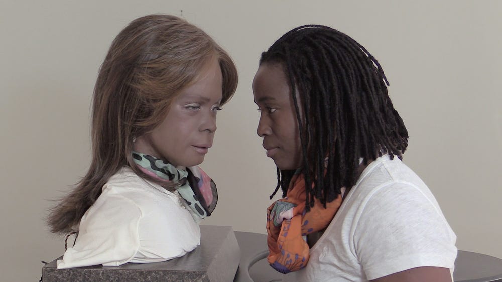Still of a Black woman face to face with a robot bust styled to resemble her