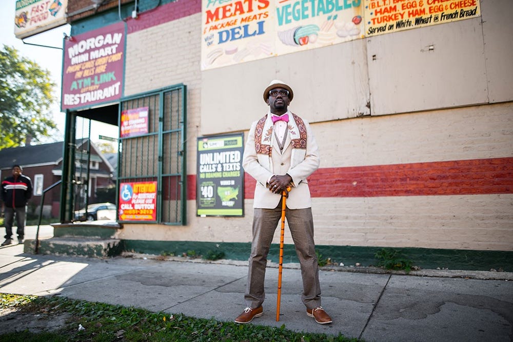 Black man in front of a market wearing a hat, shall, and bow tie, with a cane
