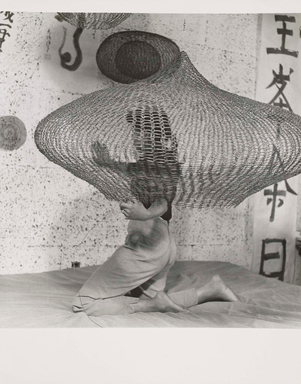 Ruth Asawa on a bed kneeling inside a looped wire sculpture