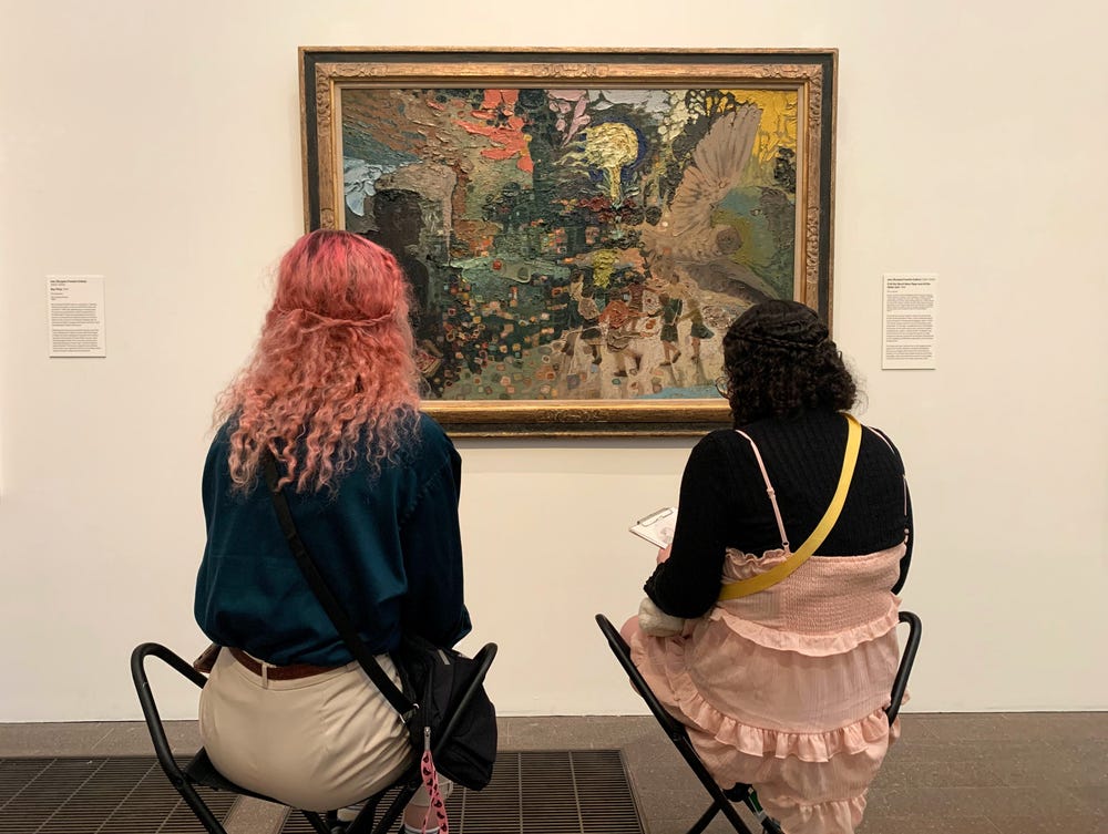 Two women, one with pink hair, sit facing a painting, sketching.