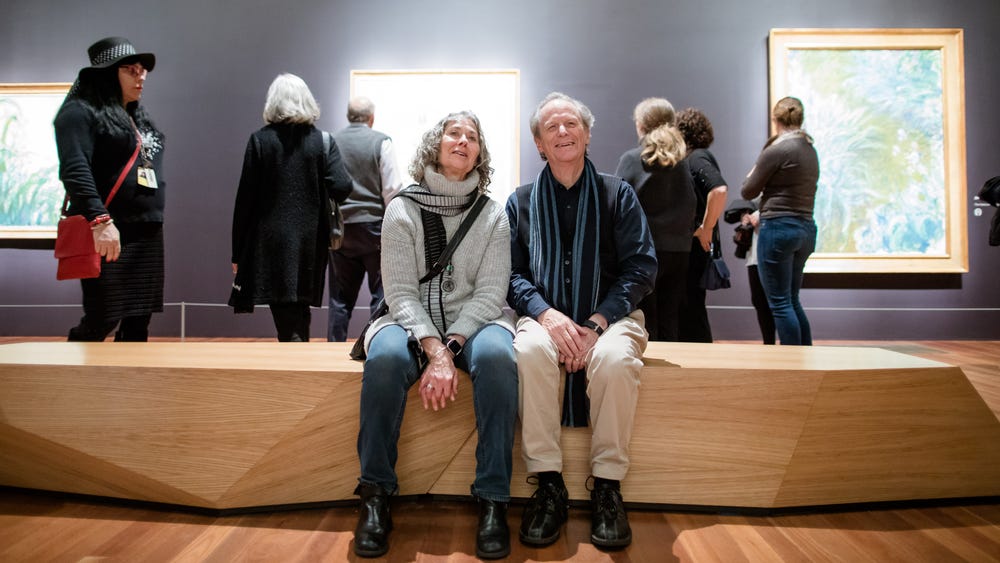 couple with gray hair sits on a bench in a gallery while people behind them look at artworks