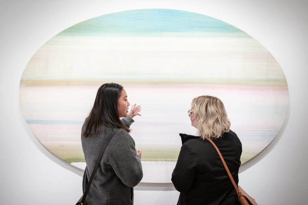 a young woman with black hair and an older woman look at an oval-shaped artwork with stripes of turquoise, white, green, and pink